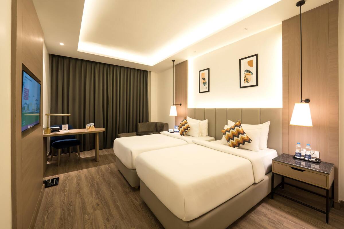 A hotel room with two beds, a desk, a chair, a TV, and a nightstand with a lamp.
