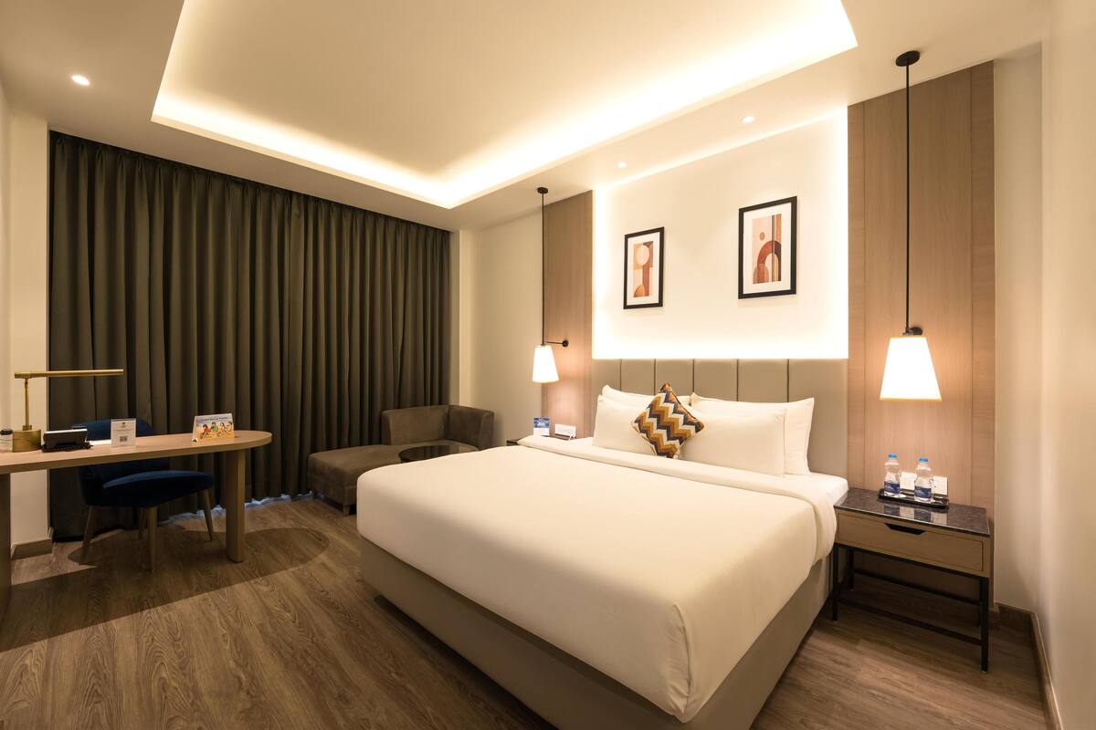 A hotel room with a large bed, a desk with a chair, a nightstand with a lamp, and two wall-mounted pictures.