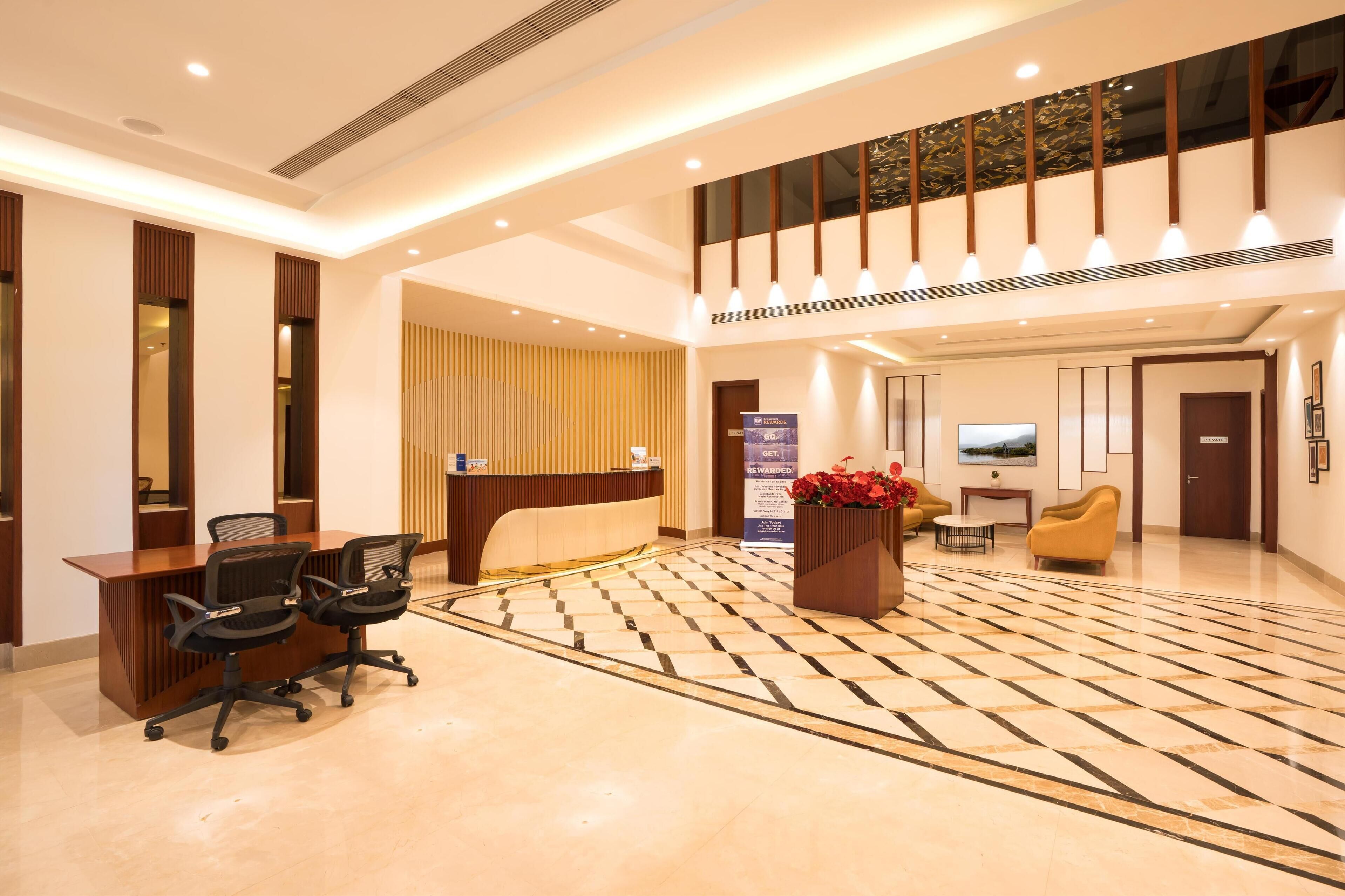A hotel lobby with a reception desk, two black office chairs, a check-in counter, a large potted plant, a television, and a patterned floor.