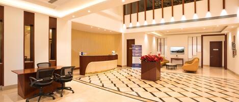 A hotel lobby with a reception desk, two black office chairs, a check-in counter, a large potted plant, a television, and a patterned floor.
