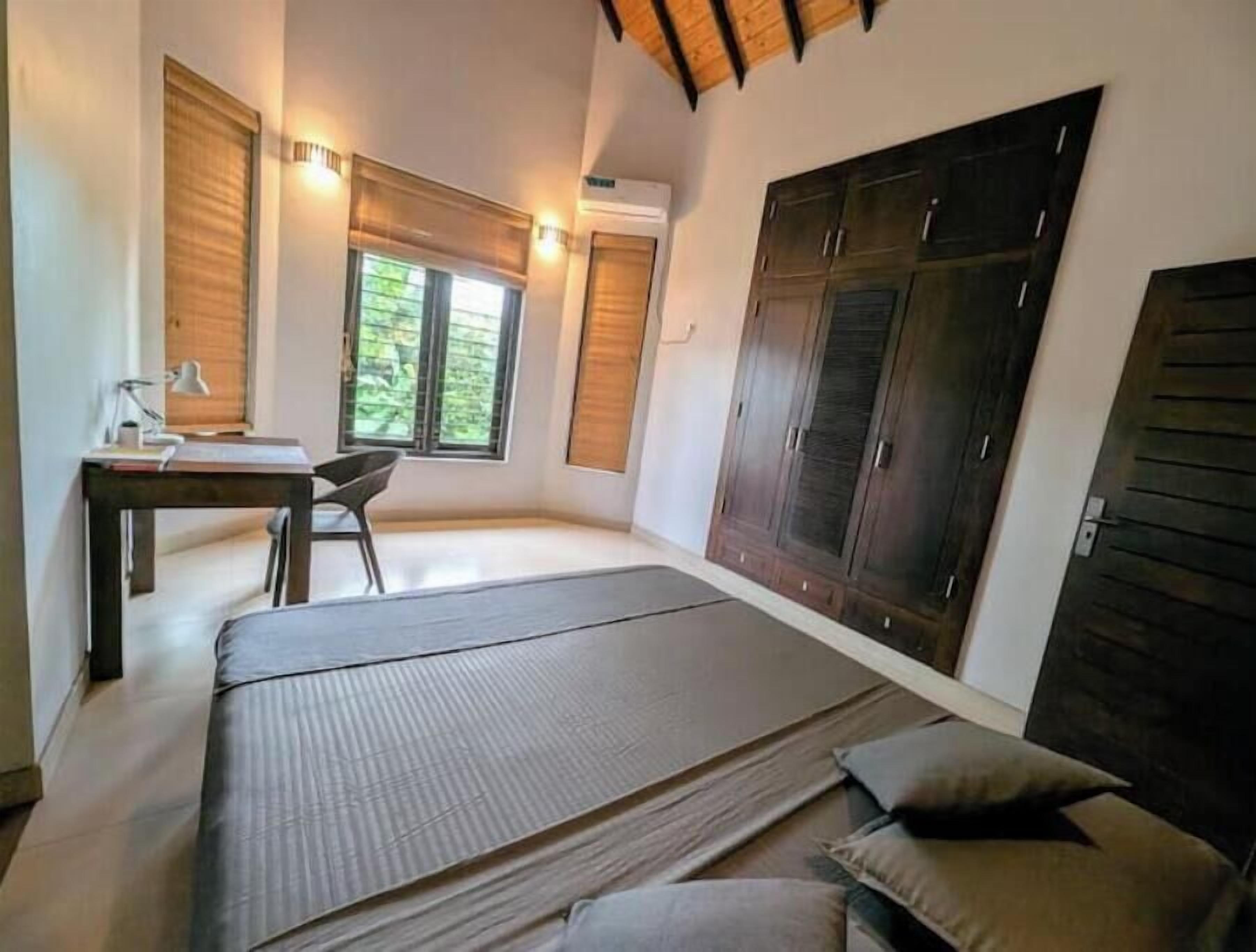 A bedroom with a bed, a desk, and a chair. There are windows with blinds, a wardrobe, and a ceiling with wooden beams.