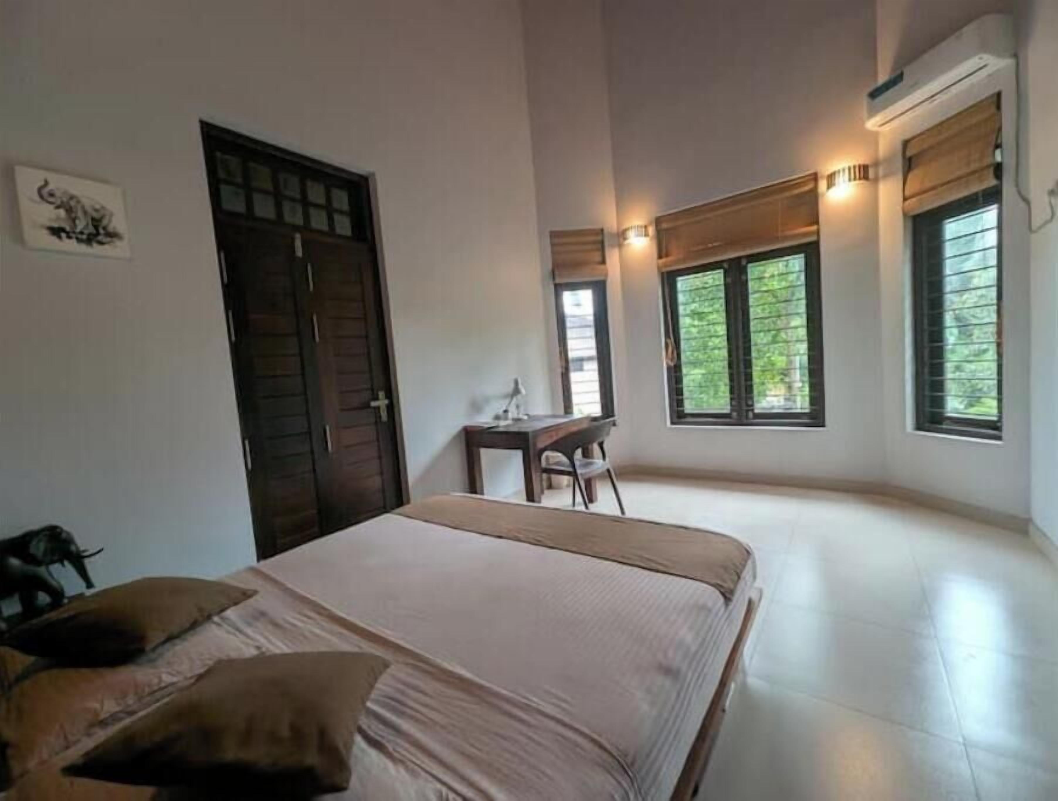 Een slaapkamer met een bed, een bureau en een stoel. Er zijn ramen met zonwering, een airconditioning en een deur.