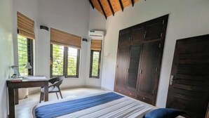 A bedroom with a bed, a desk, and a chair. There are windows with blinds, a wardrobe, and an air conditioning unit.
