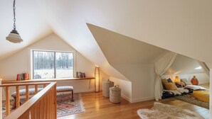 Un dormitorio acogedor en el ático con una escalera de madera, un ventanal con vistas a árboles, una zona de estar confortable con cojines y una lámpara colgante.