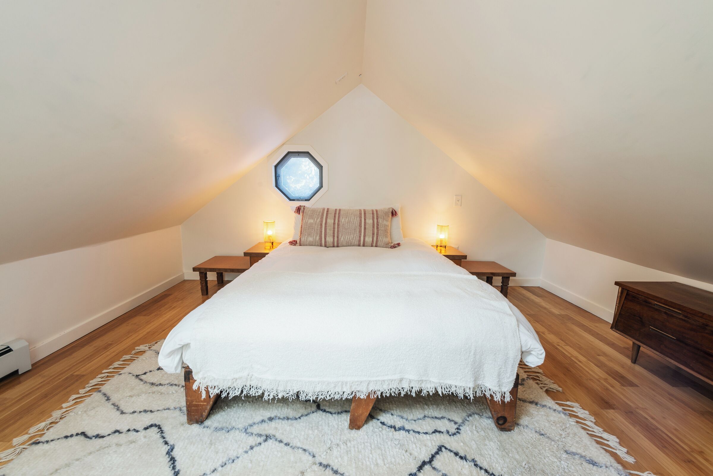 Un dormitorio con techo inclinado, una cama con manta blanca, dos mesitas de noche con lámparas, un banco de madera y una alfombra.