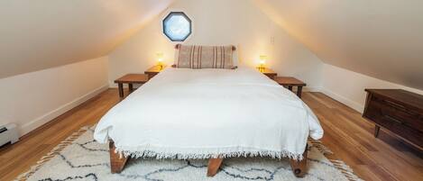 Un dormitorio con techo inclinado, una cama con manta blanca, dos mesitas de noche con lámparas, un banco de madera y una alfombra.
