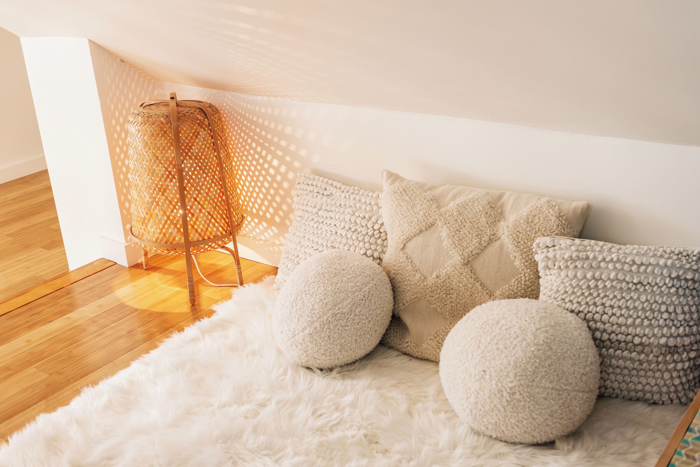 A cozy room with a white furry rug, a woven floor lamp, and several textured pillows.