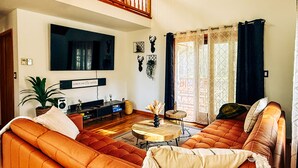 Een woonkamer met een grote oranje bank, een flatscreen televisie aan de muur, een klein rond salontafeltje en een potplant.