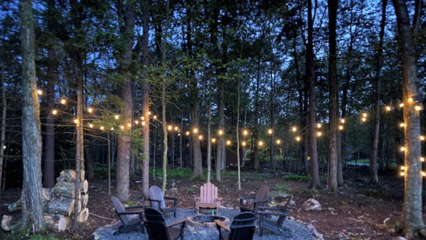 Un área de estar al aire libre con un brasero, rodeada de árboles y luces colgantes.