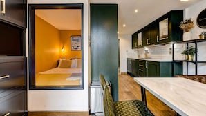 Una habitación compacta con cama, un cubo de basura pequeño, una mesa de comedor y una zona de cocina con armarios y una encimera.