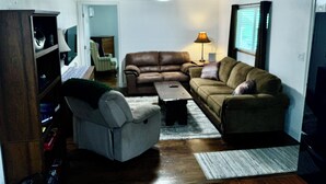 A living room with a brown leather sofa, a white armchair, a coffee table, and a lamp. There is a wooden cabinet and a television on the right.