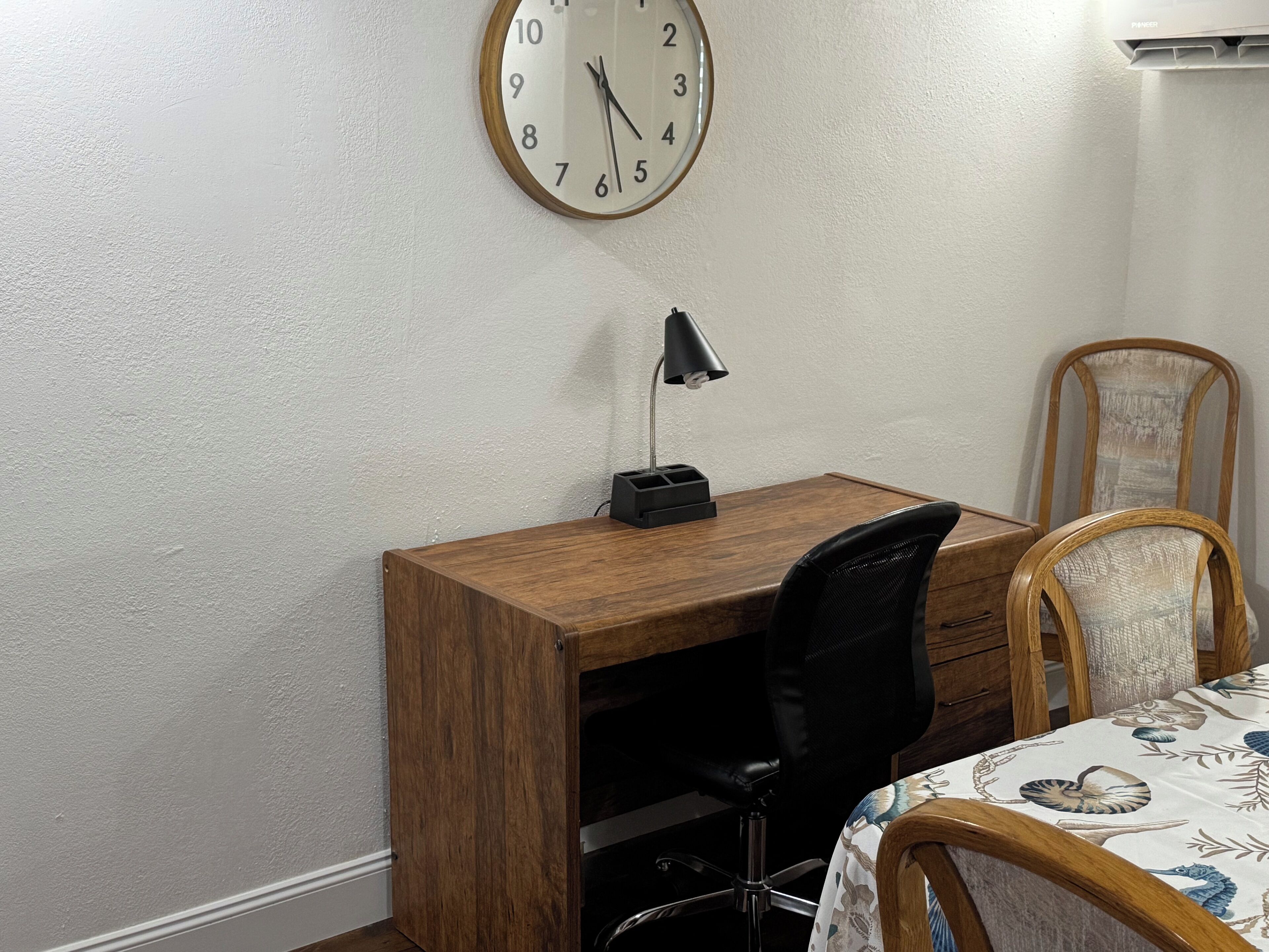 Ein Schlafzimmer mit einem Holztisch, einem schwarzen Bürostuhl, einem Bett mit floralem Überwurf und einer Wanduhr.