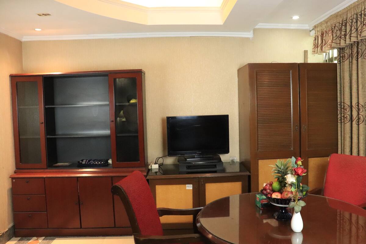 A room with a dining table, red chairs, a television on a stand, and a cabinet with glass doors.