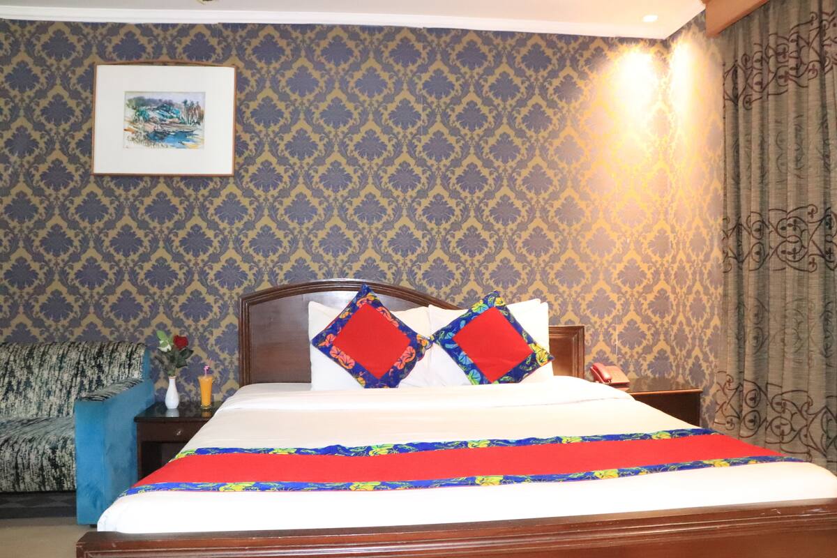 A hotel room with a bed, a nightstand, a lamp, a framed picture on the wall, and a patterned wallpaper.