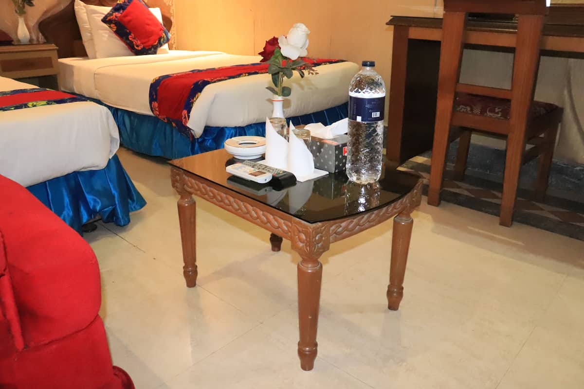 A hotel room with a bed, a glass-top table with a remote control, a water bottle, and a vase with a flower.