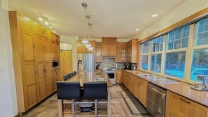 A modern kitchen with wooden cabinets, a central island with stools, stainless steel appliances, and a large window providing a view of the outdoors.