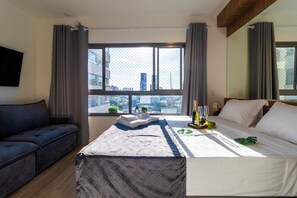 A modern hotel room with a large window, a bed, a sofa, and a small table with amenities. - Modern, fully equipped, and well-lit with AC and gym (Sao Paulo)