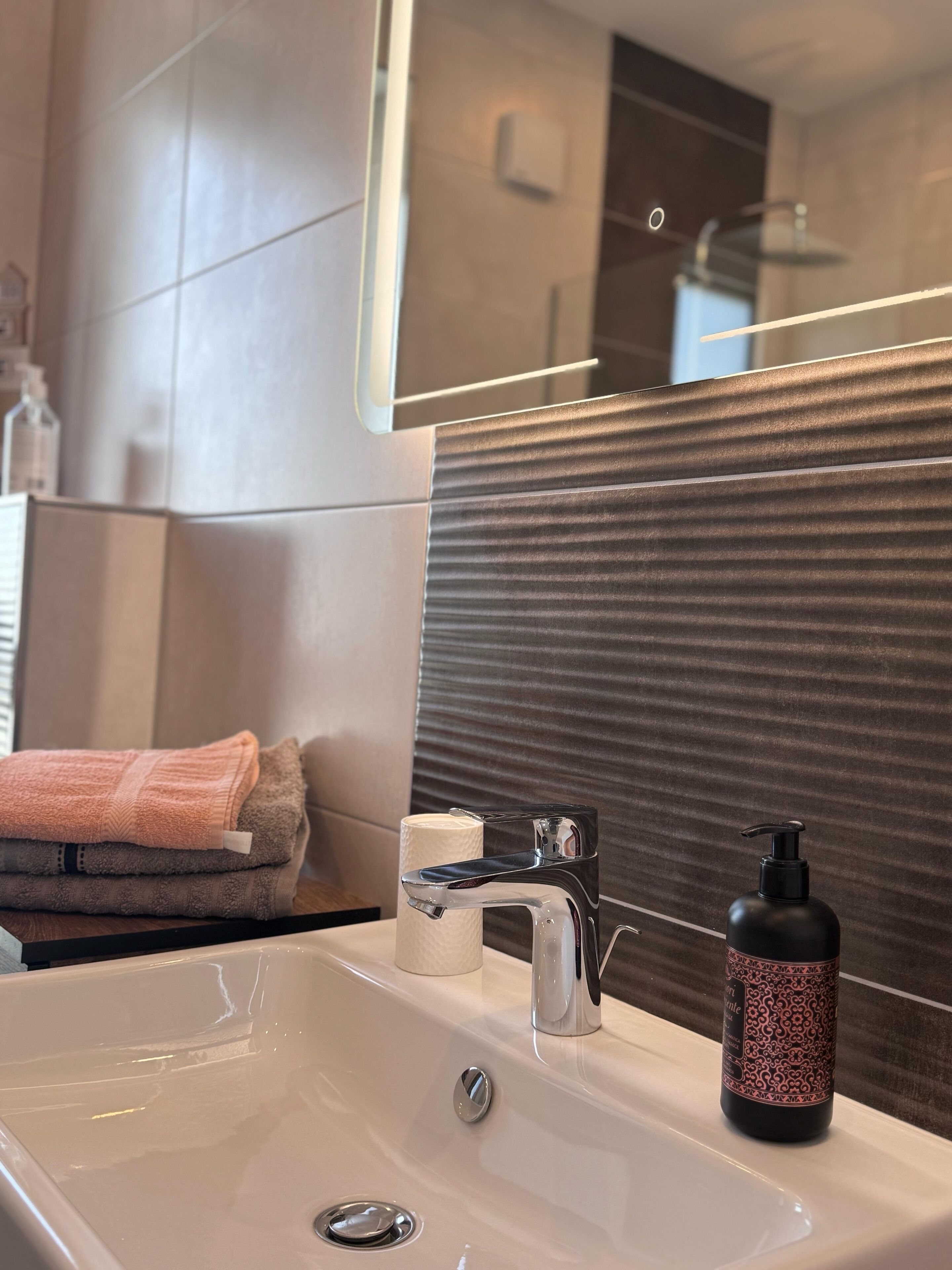 A modern bathroom with a large mirror, a white sink, a chrome faucet, and a bottle of shampoo.
