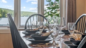 Una mesa de comedor dispuesta con platos negros, cuencos y copas de vino transparentes, ubicada junto a una ventana con vista a un lago y follaje.