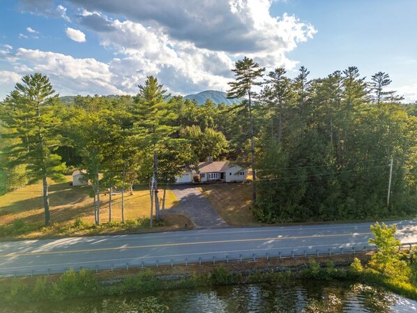 A house with a driveway leading to a road, surrounded by trees and a body of water.