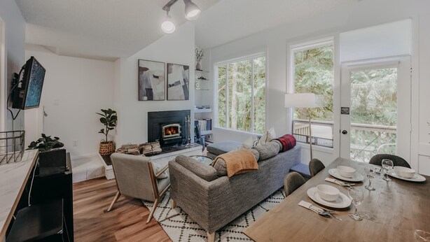 Ein modernes Wohnzimmer mit Kamin, Sofa, Esstisch und großen Fenstern mit Blick auf Bäume.