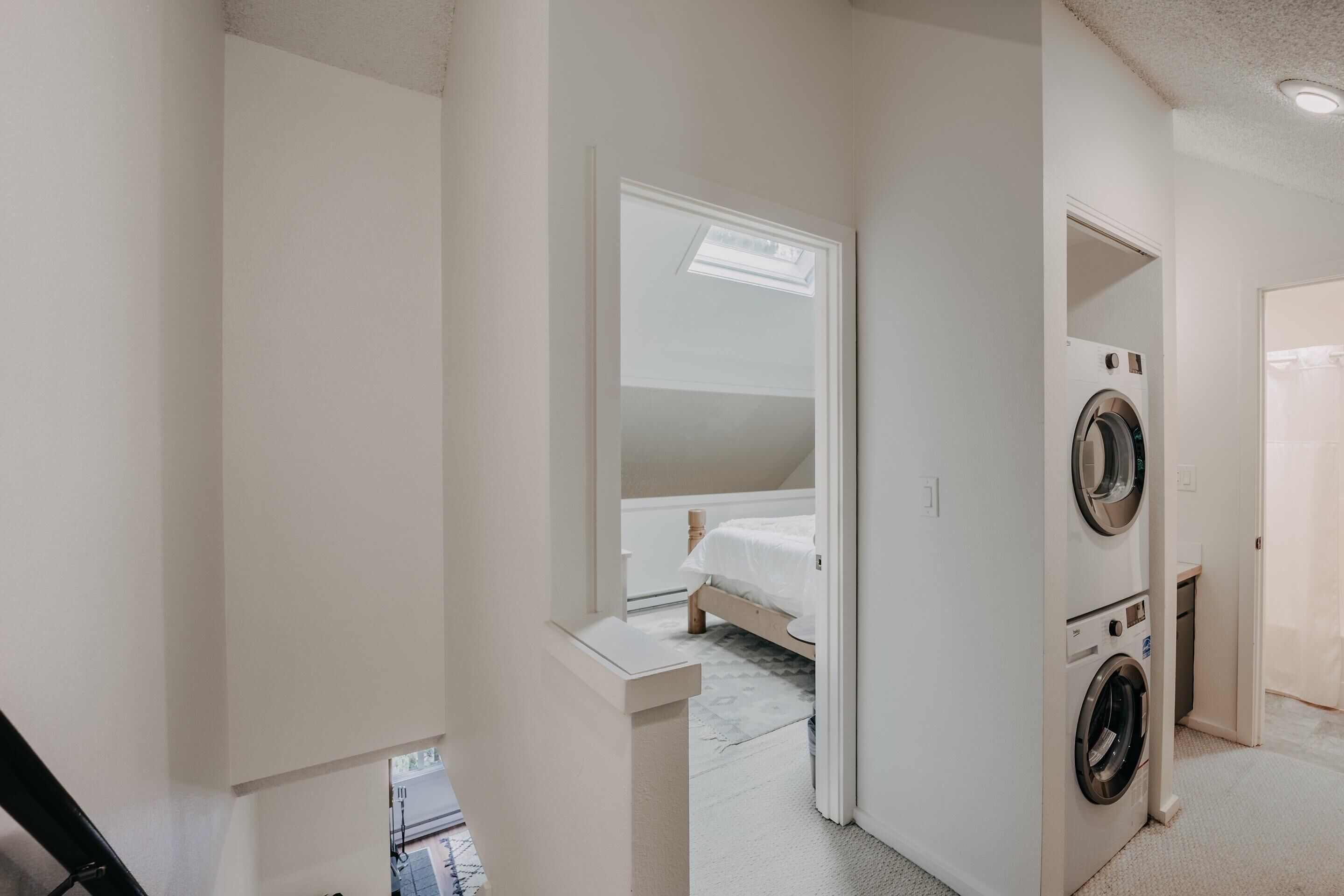 A laundry room with two washing machines, a bed in the adjacent room, and a skylight above the bed.