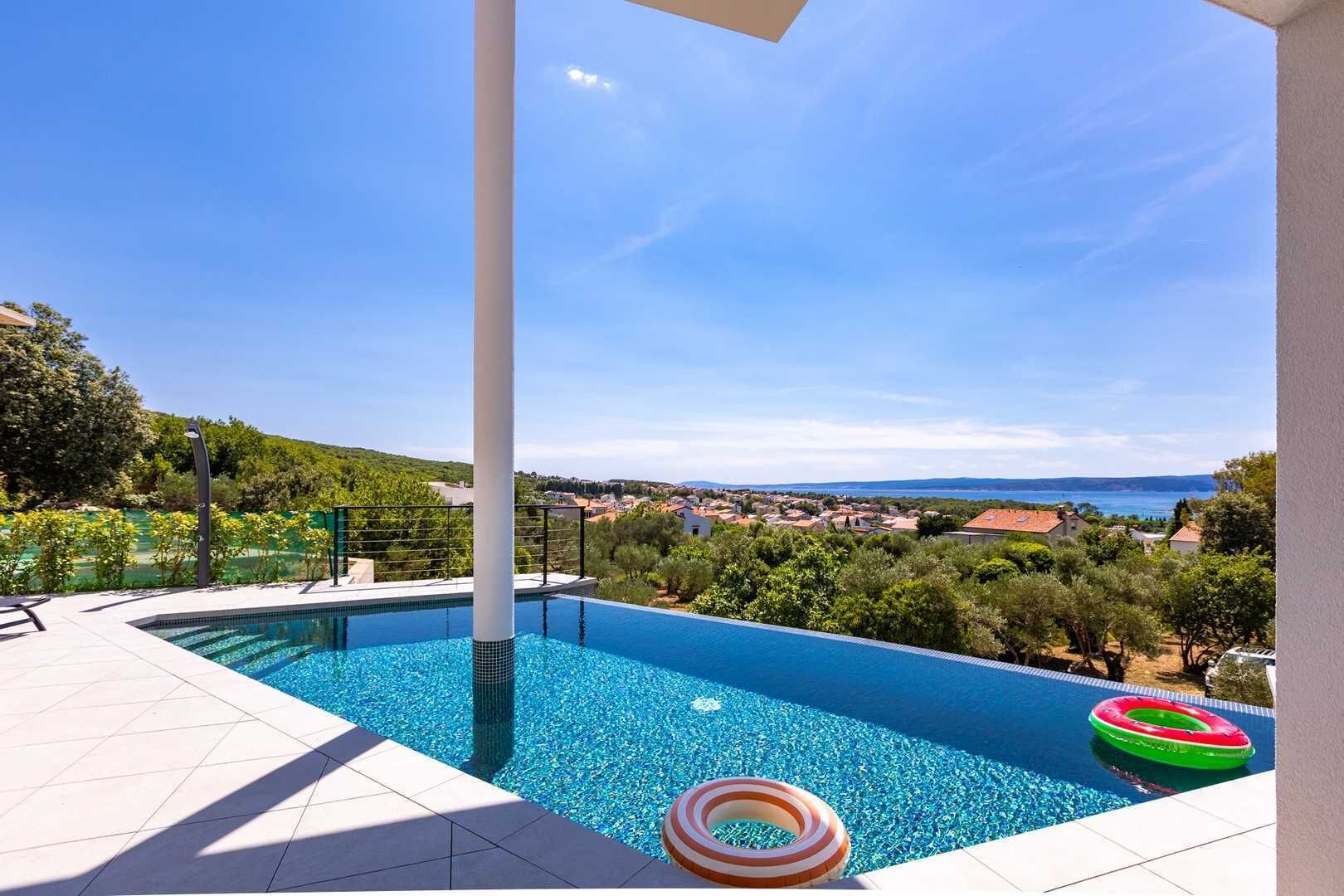 A swimming pool with clear blue water, surrounded by a white tiled deck. There are two inflatable rings on the poolside. The pool is set against a backdrop of a clear sky and a distant view of a town and coastline.