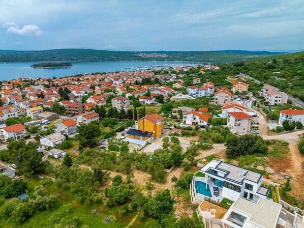 Vue aérienne d’une zone résidentielle côtière, avec des maisons, une marina et un paysage vallonné en arrière-plan.