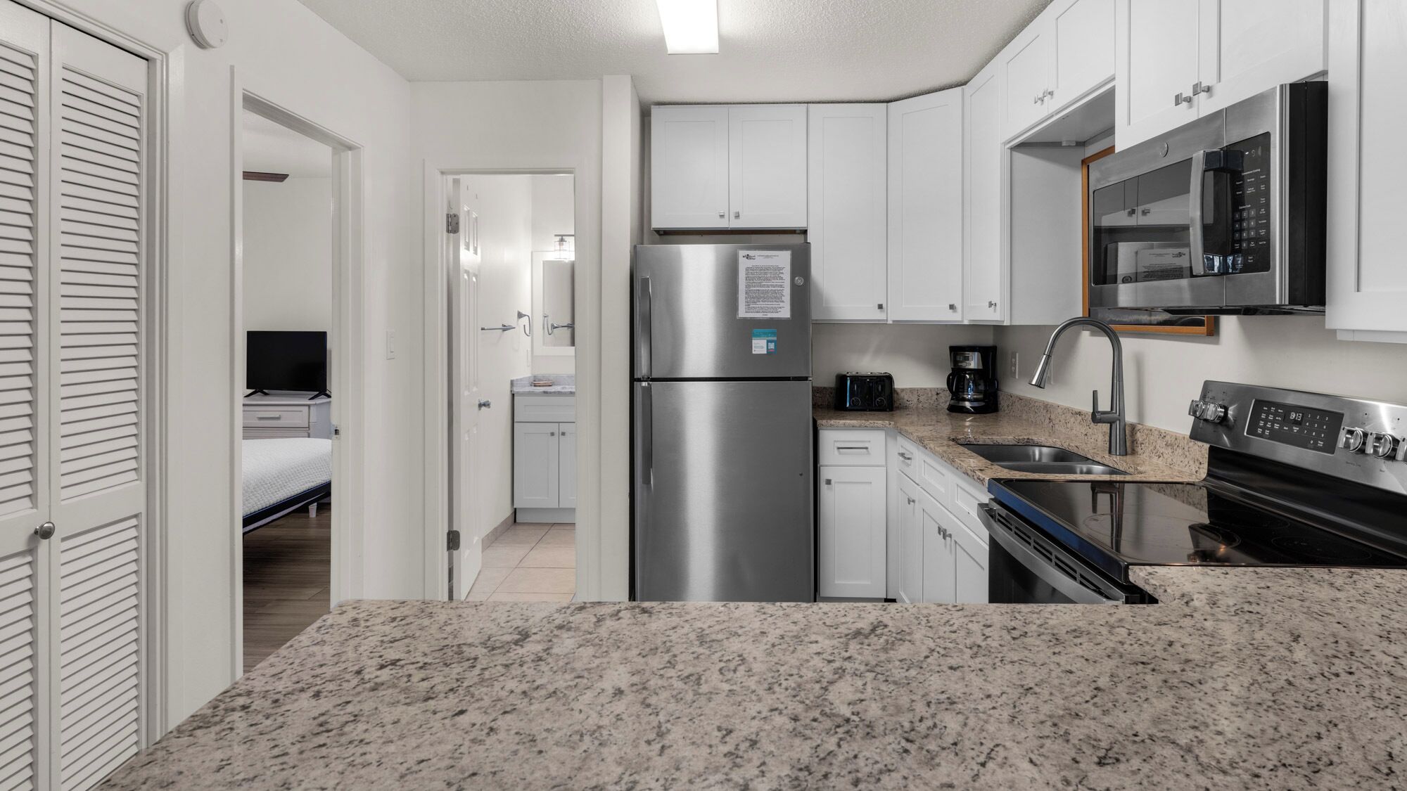 A compact kitchen with white cabinets, a stainless steel refrigerator, a microwave, a coffee maker, and a stove.
