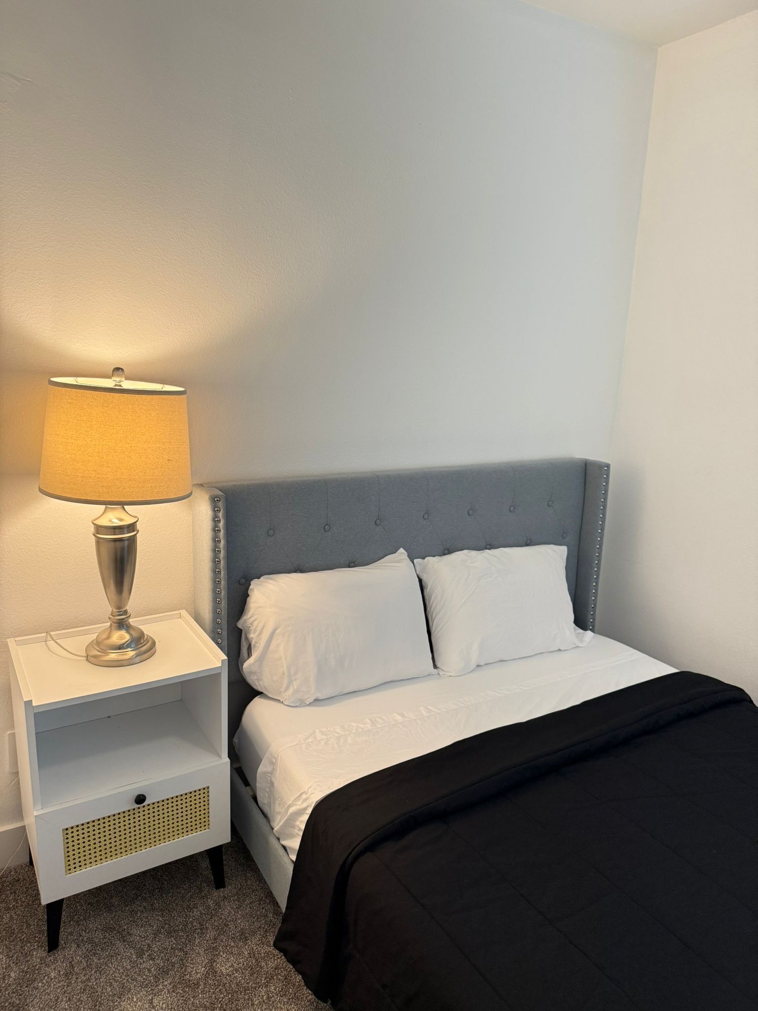 A neatly made bed with white linens and a black blanket, a grey upholstered headboard, and a bedside table with a lamp.