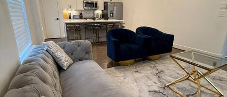 A modern living room with a grey sectional sofa, a blue armchair, and a glass coffee table with a gold frame.