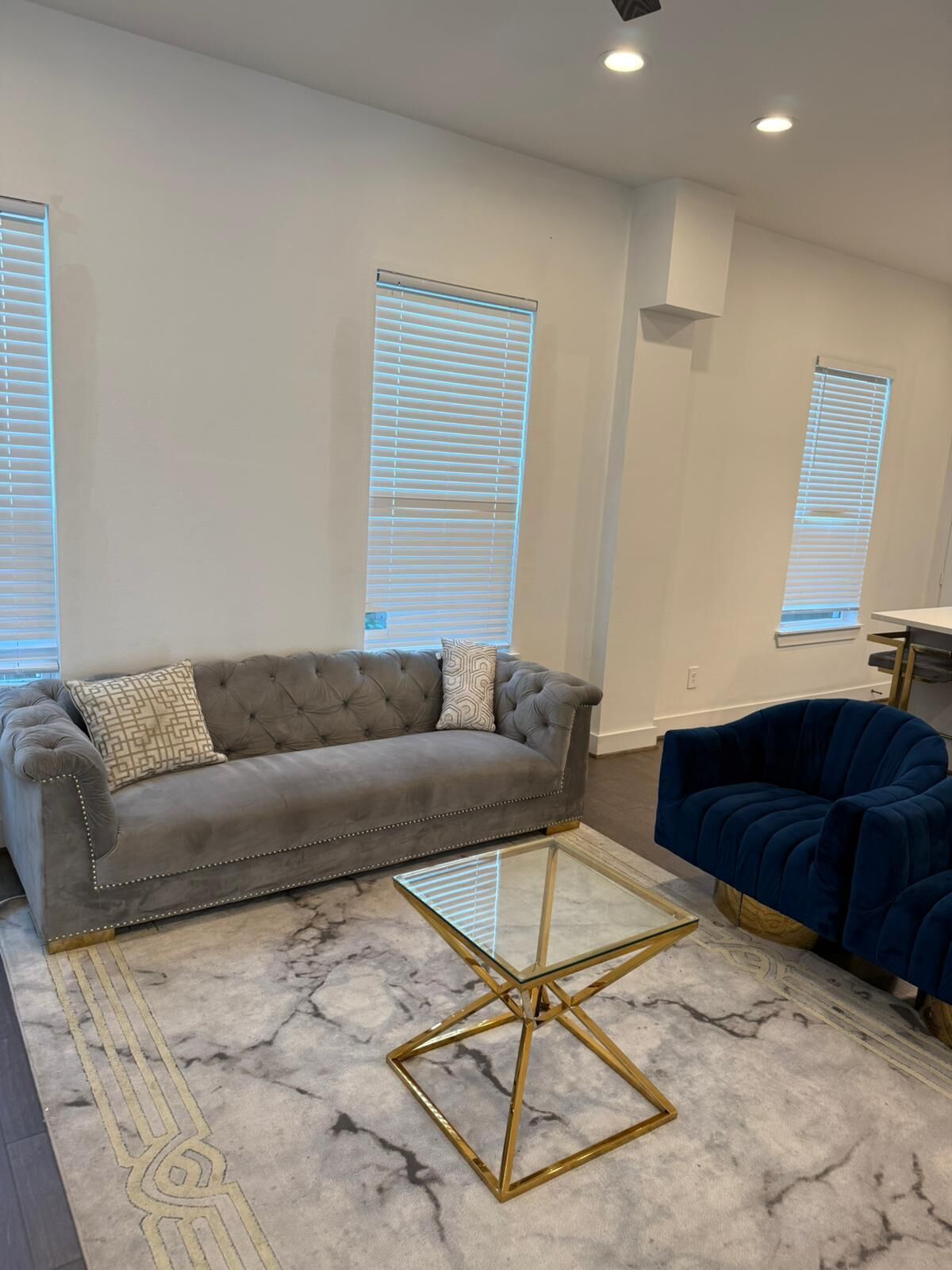 A living room with a grey tufted sofa, a blue velvet sofa, and a glass coffee table with a gold frame.