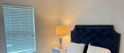 Een slaapkamer met een donkerblauwe bed, witte kussens, een grijze deken, een bedlampje en een plafondventilator.