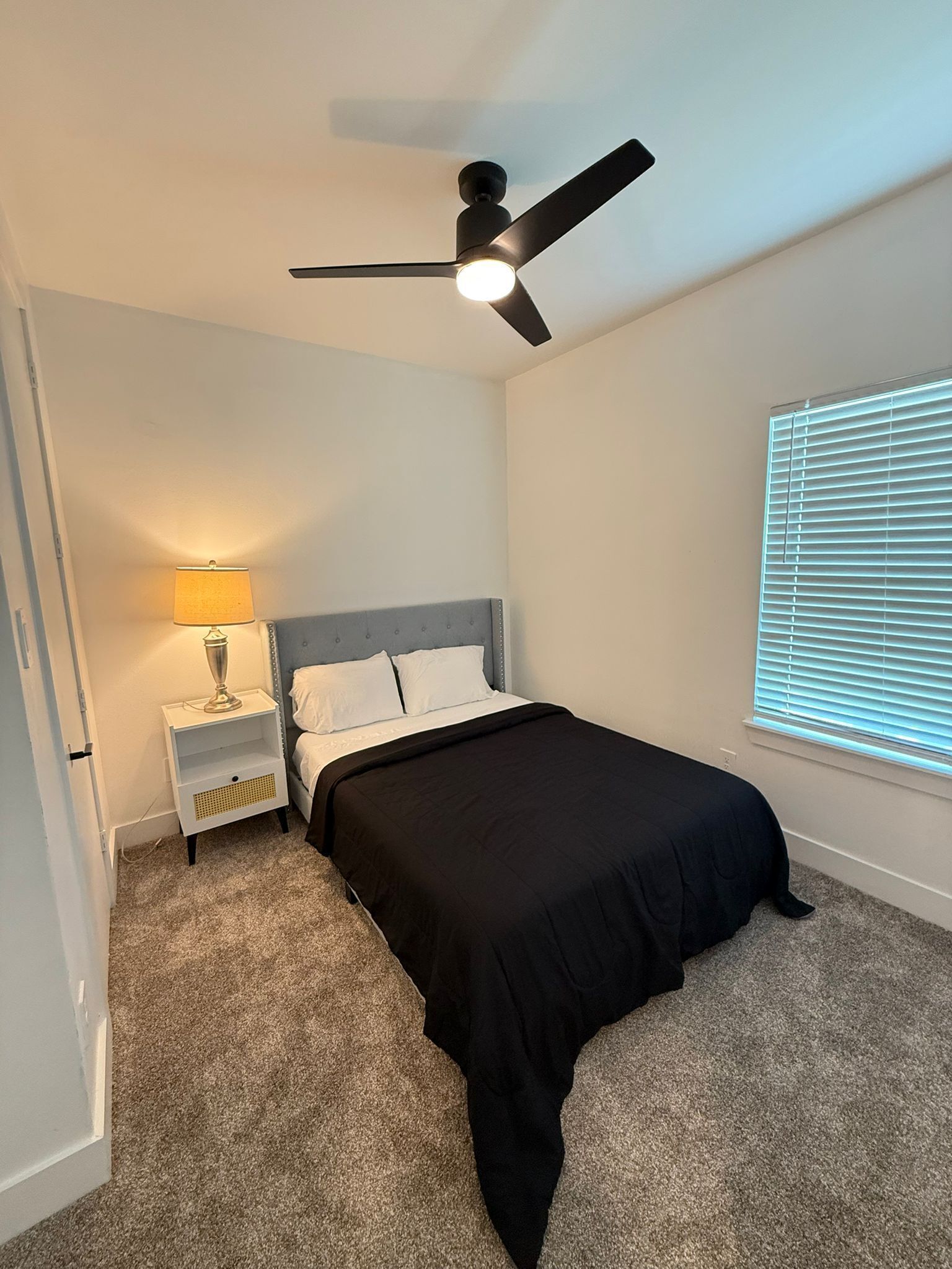 A bedroom with a bed, a nightstand, a lamp, a ceiling fan, and a window with blinds.