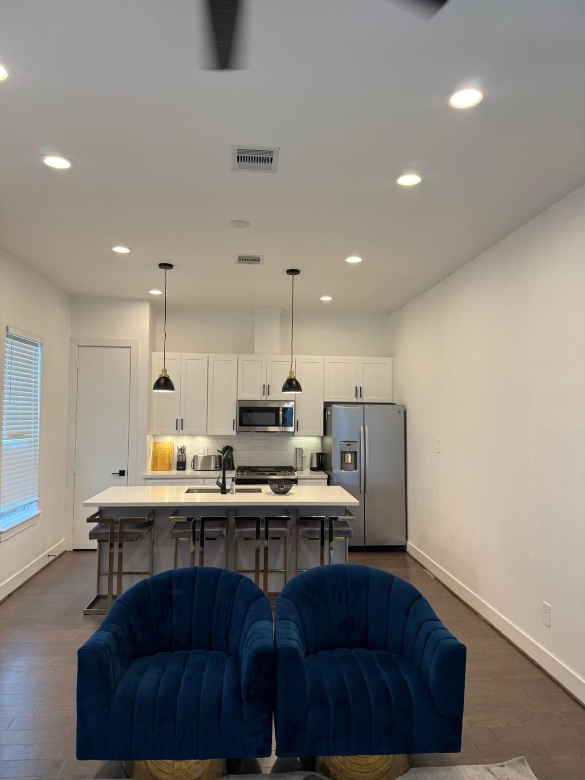 Una cucina moderna con mobili bianchi, elettrodomestici in acciaio inossidabile e illuminazione a sospensione. In primo piano, due poltrone blu.