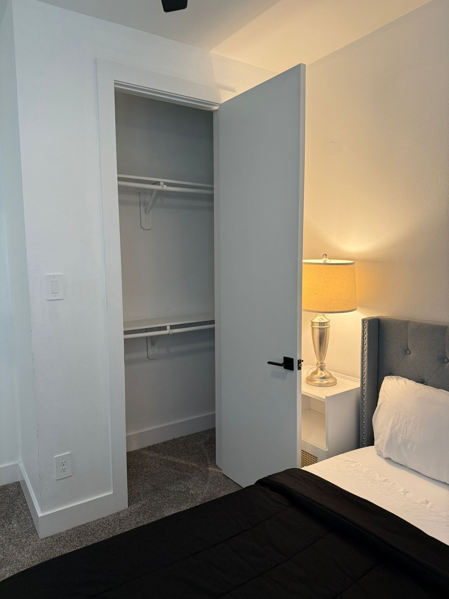 A hotel room with a bed, a bedside lamp, a nightstand, and a closet with hanging clothes.