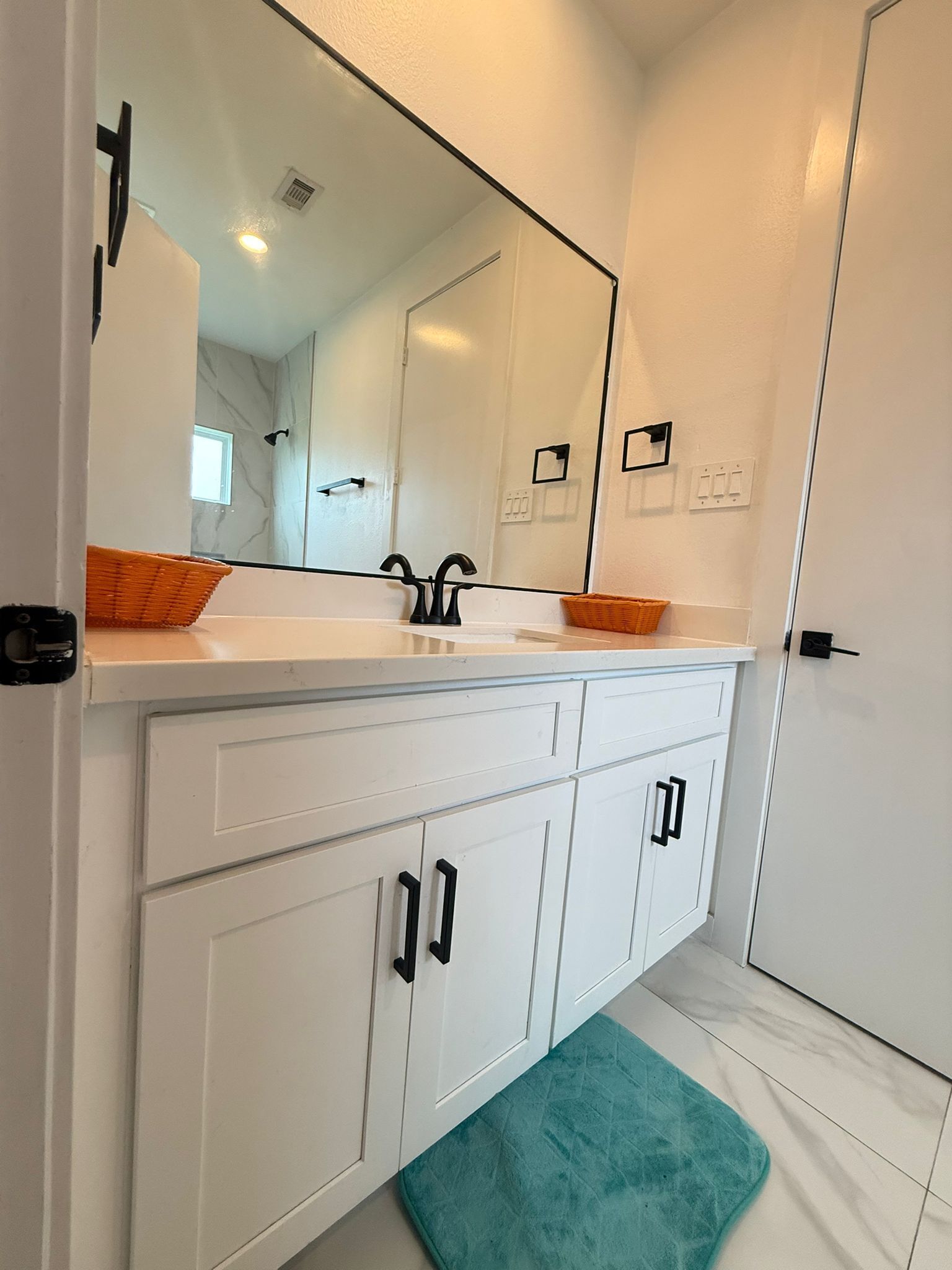 A bathroom with a white vanity, two sinks, a large mirror, and a teal bath mat.