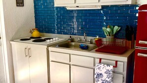 Una cocina compacta con armarios blancos, salpicadero de azulejos azules, hervidor amarillo, un lavabo y un refrigerador rojo.