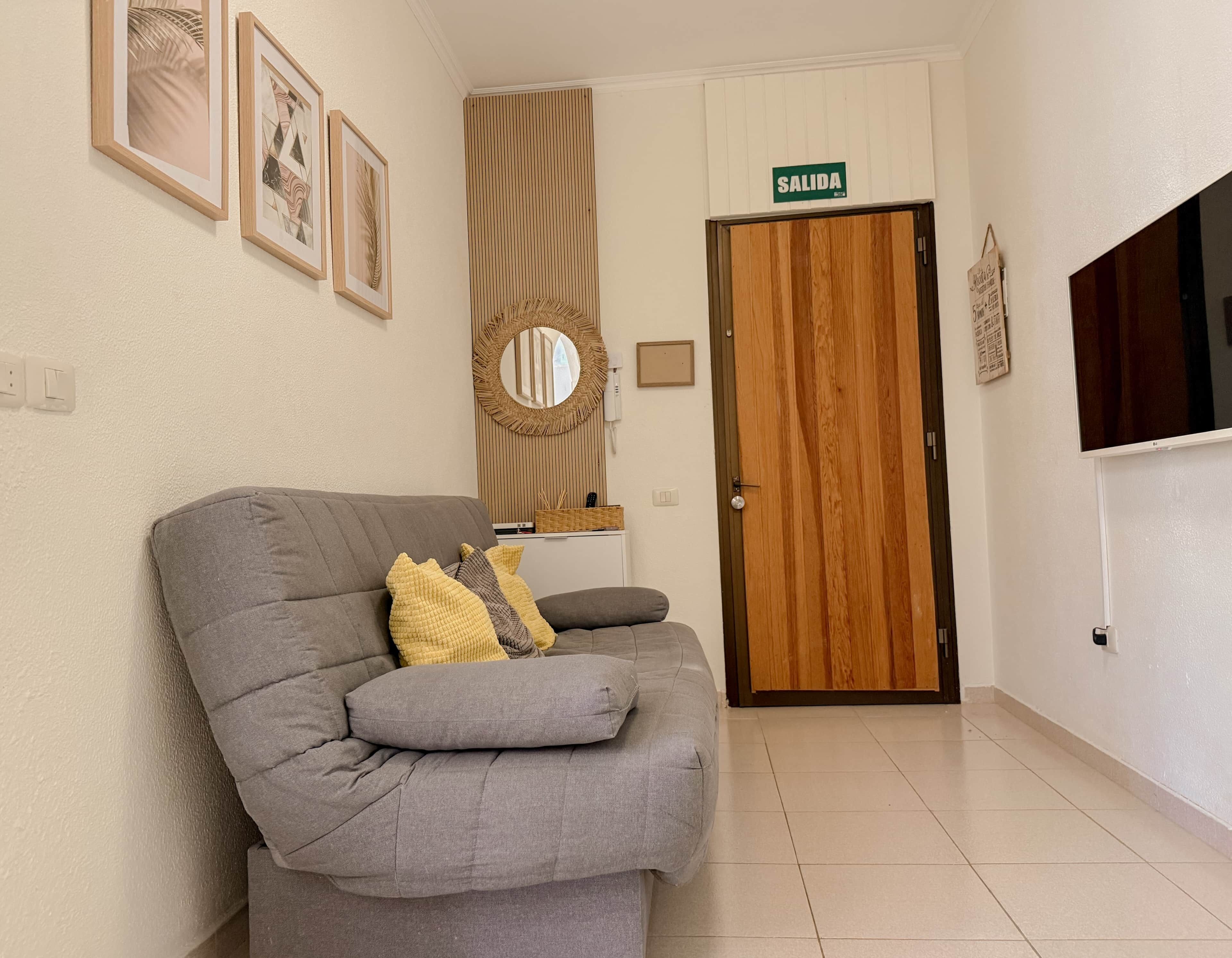 A living room with a grey sofa, framed artwork, a mirror, and a 'SALIDA' sign above a wooden door.