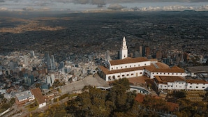 An aerial view of a city with a prominent white building with a tower, surrounded by dense urban development and a distant mountain range. - Kaza Living Calle 100 (Bogotá)