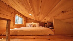 A cozy bedroom with a wooden ceiling, a window, a bed with white bedding, and a small bookshelf.