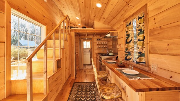 A wooden cabin interior with a kitchen area, a dining table, and a staircase leading to an upper level.