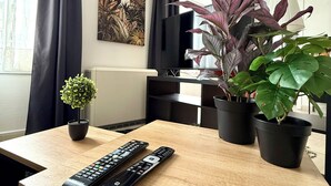 Een woonkamer met een houten tafel, twee afstandsbedieningen, een potplant, een televisie en een schilderij aan de muur.