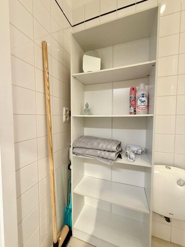 A bathroom with white tiled walls, a wooden bath mat leaning against the wall, a shelf with toiletries, and a white toilet. - Peaceful 2 minutes European Parliament (Brussels)