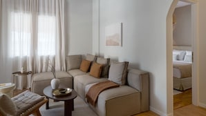 Ein modernes Wohnzimmer mit einem beigen Sofa, einem kleinen runden Tisch und einem Sessel. Ein Fenster mit transparenten Vorhängen bietet Blick ins Freie.