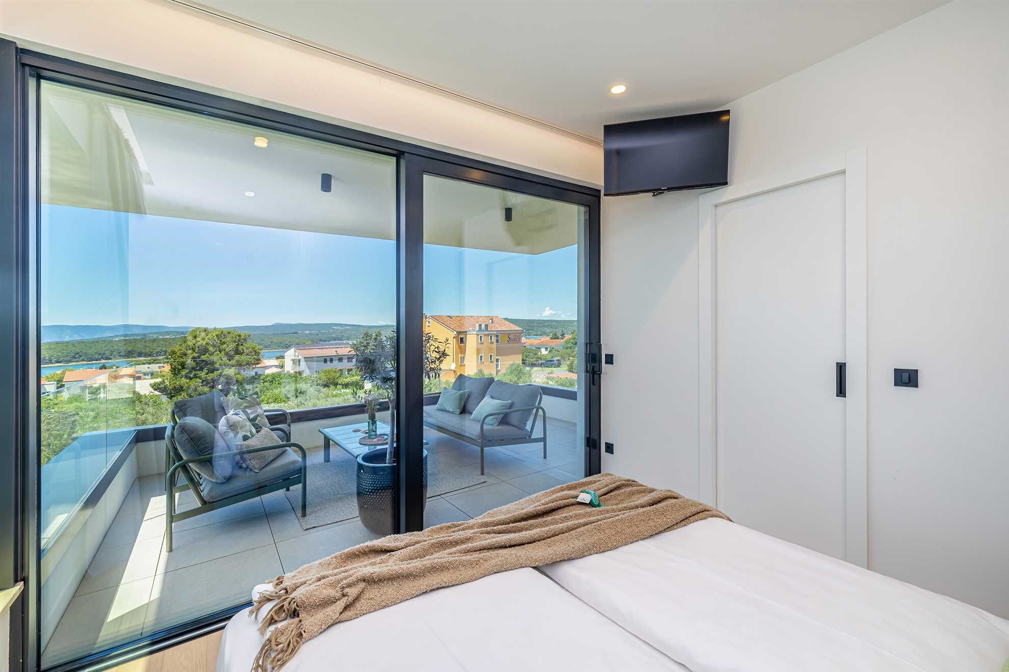 A bedroom with a large sliding glass door leading to a balcony with a sofa and table, a bed with a blanket, and a TV mounted on the wall.