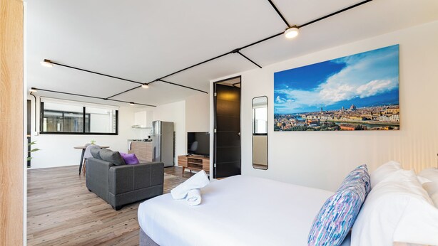 A modern hotel room with a large bed, a sofa, a TV, and a cityscape painting.