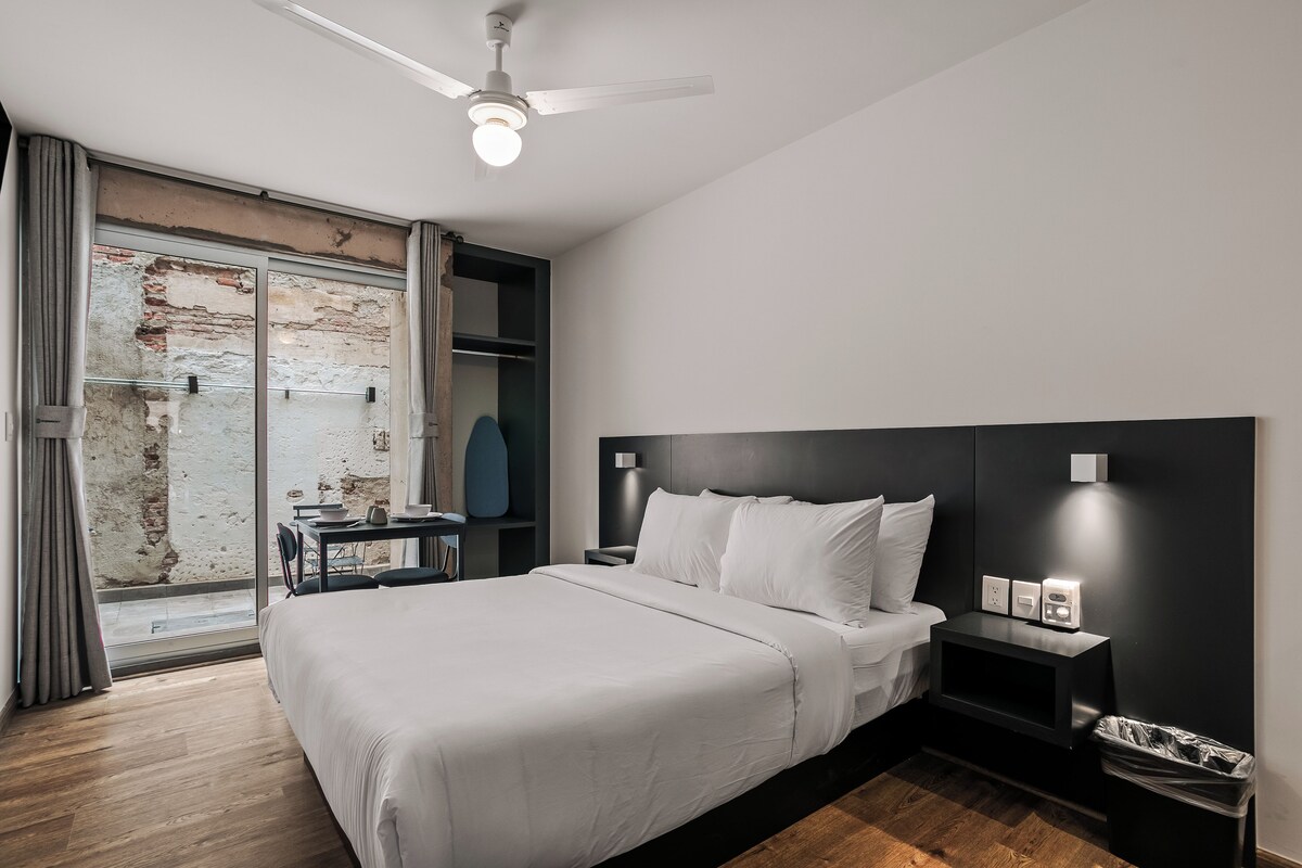 A hotel room with a bed, bedside table, nightstand, and a view of a brick wall through a glass door.