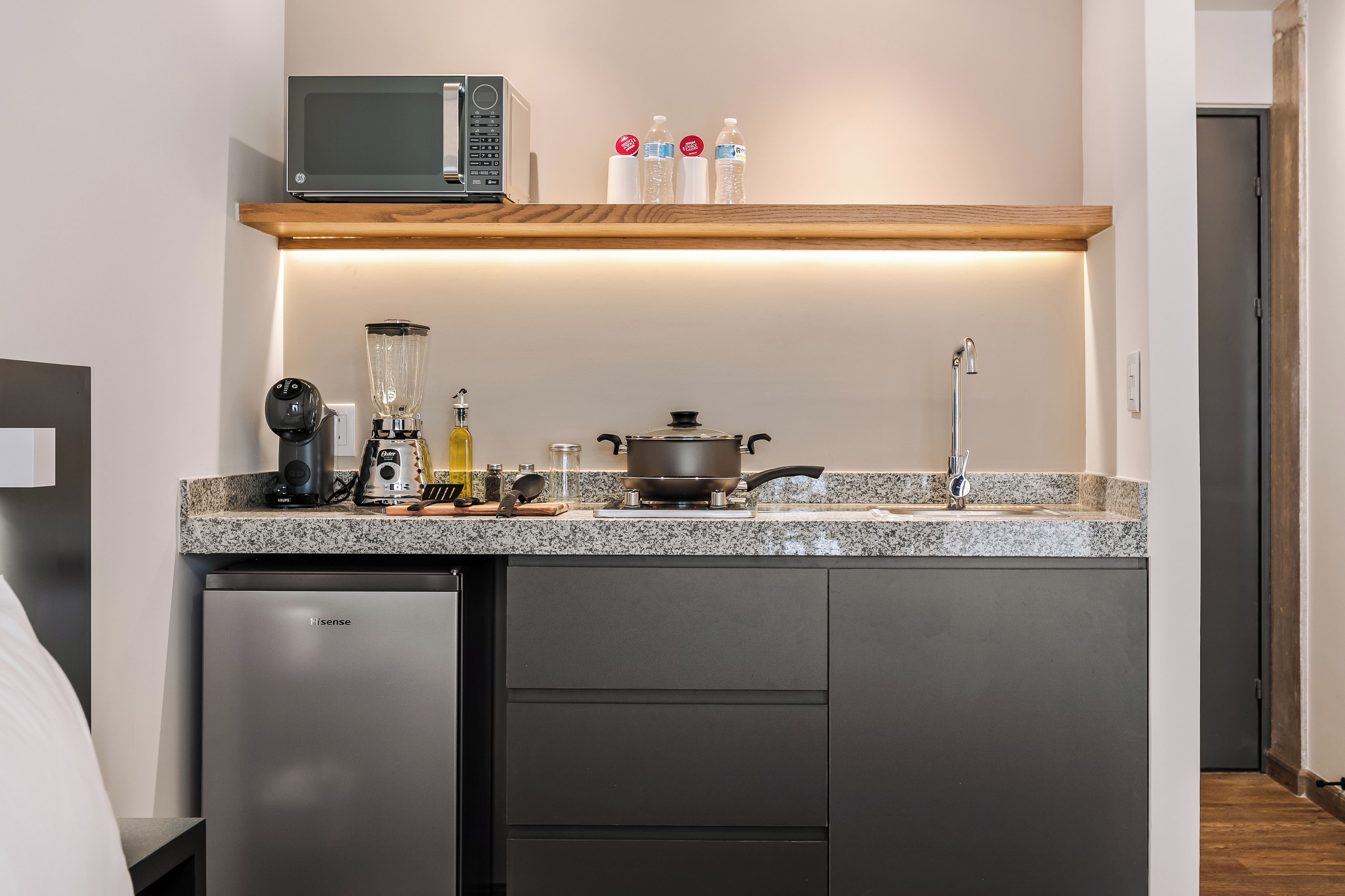 A compact kitchen with a microwave, a blender, a kettle, and a small refrigerator.