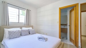A bedroom with a bed, a window with curtains, a bathroom visible through an open door, and a tiled floor.